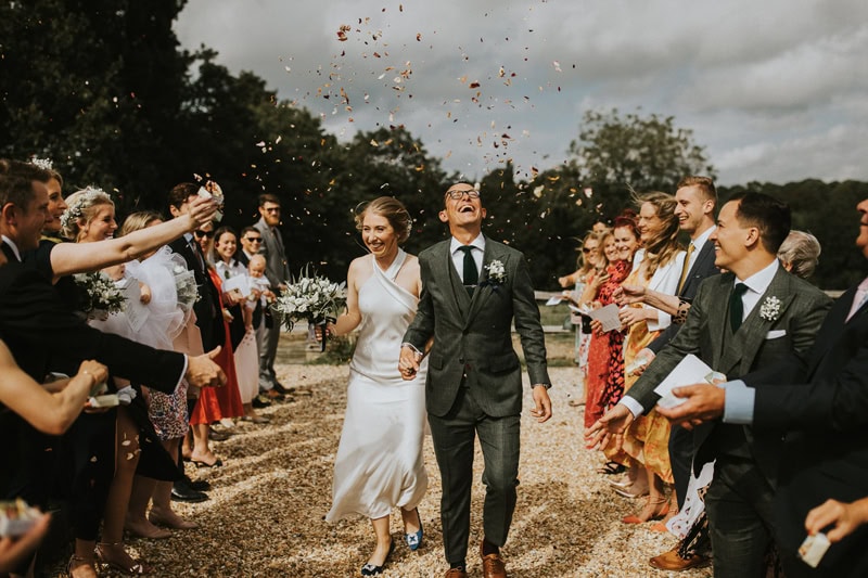 Kent Wedding Photographer, Bride and Groom walk outside on surrounded by friends and family, flowers float down around them, the forest behind them