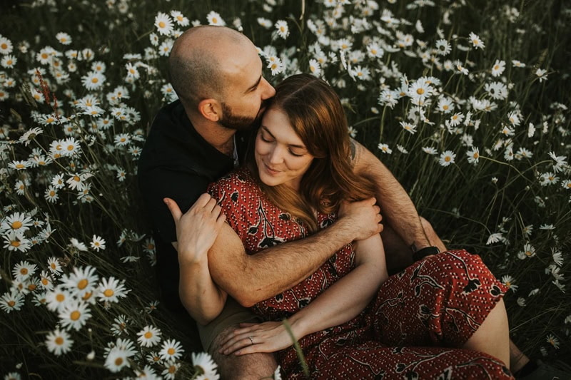 Portrait Photographer, a husband holds and kisses his wife on the head while they sit closely in the wildflowers