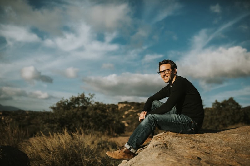 Kent Wedding Photography, Photographer Peter Reynolds of Kent, UK sits on a boulder in the outdoors with his camera, He smiles with blue sky and scattered clouds behind him