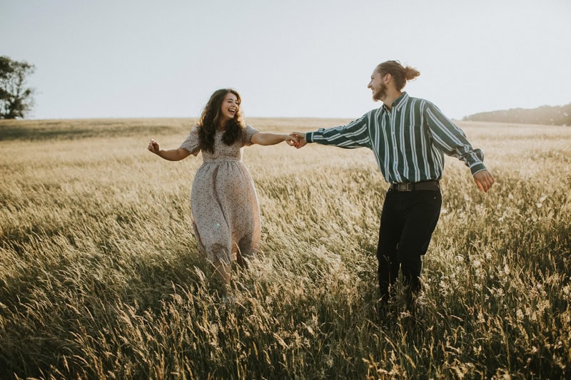 A couple joyfully running hand in hand through a sunlit grassy field, both laughing and wearing casual outfits. The scene captures a playful and carefree moment in nature.