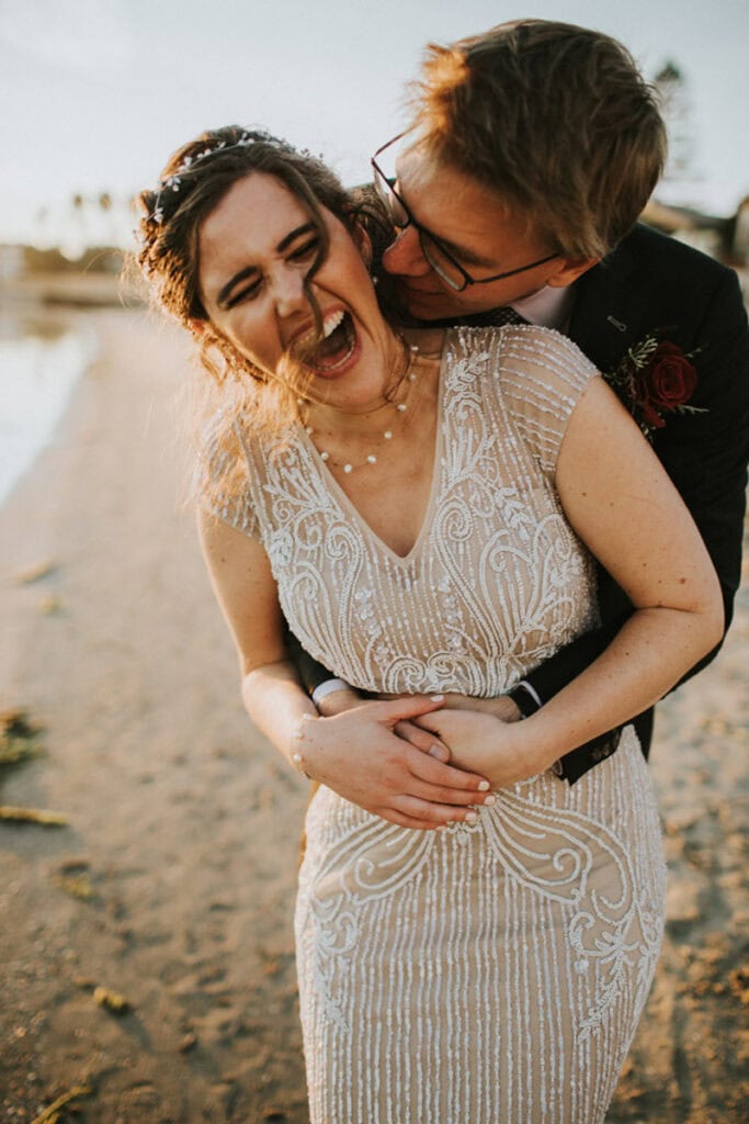 A bride laughing joyfully as her groom playfully kisses her neck while embracing her from behind. They are standing on a sandy beach during golden hour, with the bride wearing a detailed beaded gown and the groom in a suit.