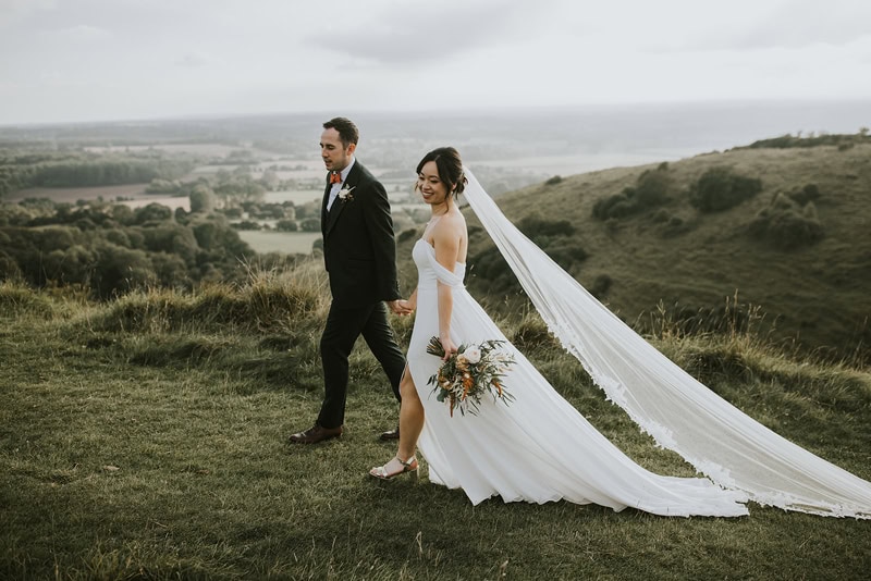 A bride and groom walking hand in hand on a grassy hill with a sweeping countryside view behind them. The bride's long veil flows in the wind, and she is holding a bouquet of flowers while the groom walks beside her in a suit.