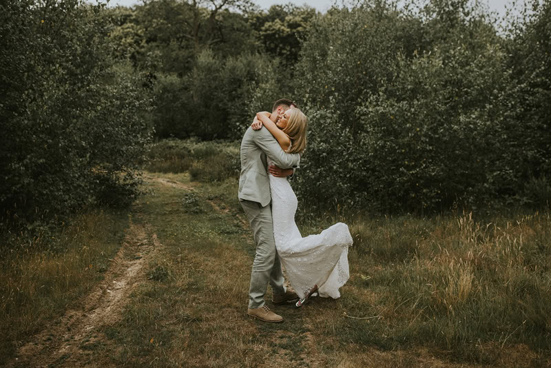 A couple embracing tightly on a dirt path in a forested area. The bride is wearing a white lace wedding dress, and the groom is dressed in a light-colored suit. They are wrapped in a warm, intimate hug surrounded by trees.