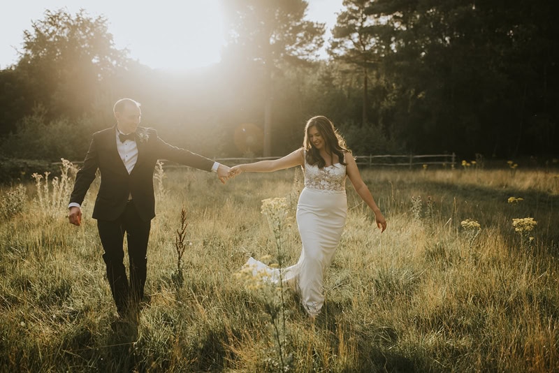 A bride and groom walking hand in hand through a sunlit field of wildflowers. The bride wears a simple, elegant wedding gown, and the groom is dressed in a tuxedo, with the warm glow of the setting sun illuminating the scene.